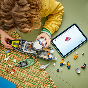 Child assembling LEGO Police Boat Chase set 60456 with yellow jacket and green wristband, surrounded by LEGO bricks, minifigures and boat pieces on green surface with tablet showing instruction app