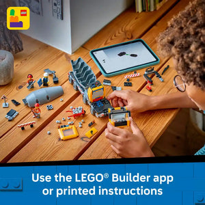 Child's hands assembling a yellow and blue LEGO cement mixer truck on a wooden table with instruction manual displayed on tablet, surrounded by LEGO bricks, minifigures and construction accessories