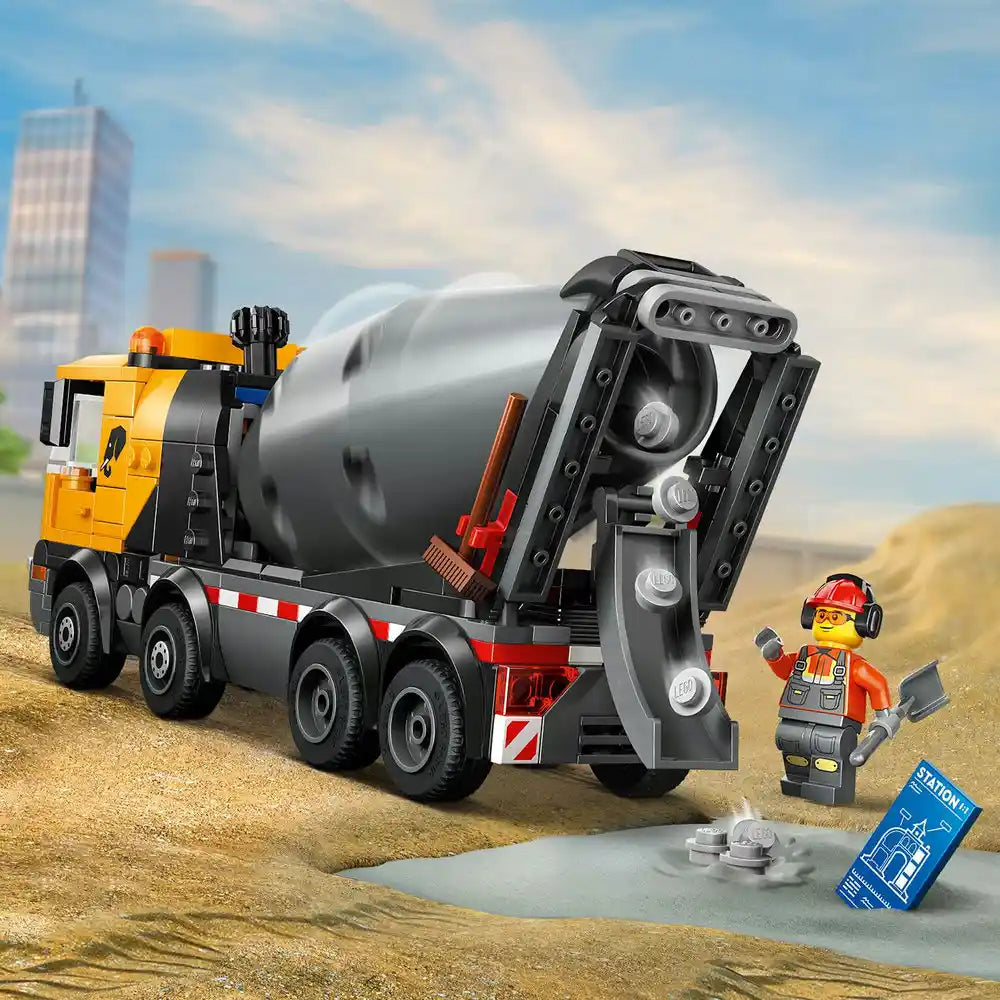 LEGO City cement mixer truck set 60478 featuring yellow and black construction vehicle with rotating gray cement drum, red and white striped details, worker minifigure in orange safety gear, and construction site accessories on sandy terrain with cityscape background