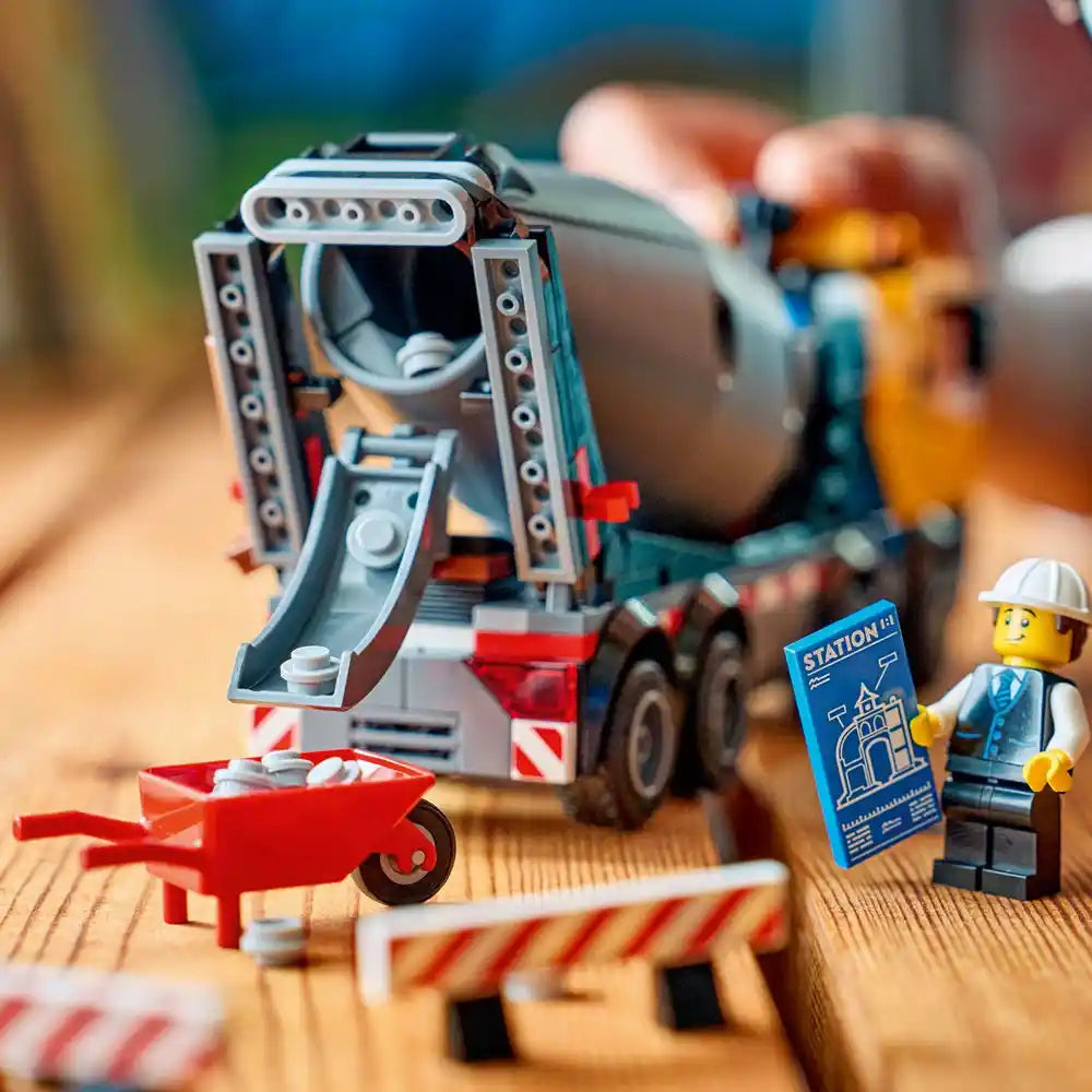 LEGO Cement Mixer 60478 construction set featuring a gray and red cement mixer truck with rotating drum, minifigure construction worker in hard hat holding blue instruction manual, red wheelbarrow, and white and red striped barrier on wooden display surface