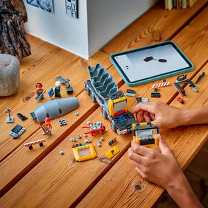 Child's hands assembling LEGO Cement Mixer 60478 set on wooden floor, displaying yellow and black construction vehicle with minifigures, building instructions on teal tablet, and scattered LEGO pieces and accessories
