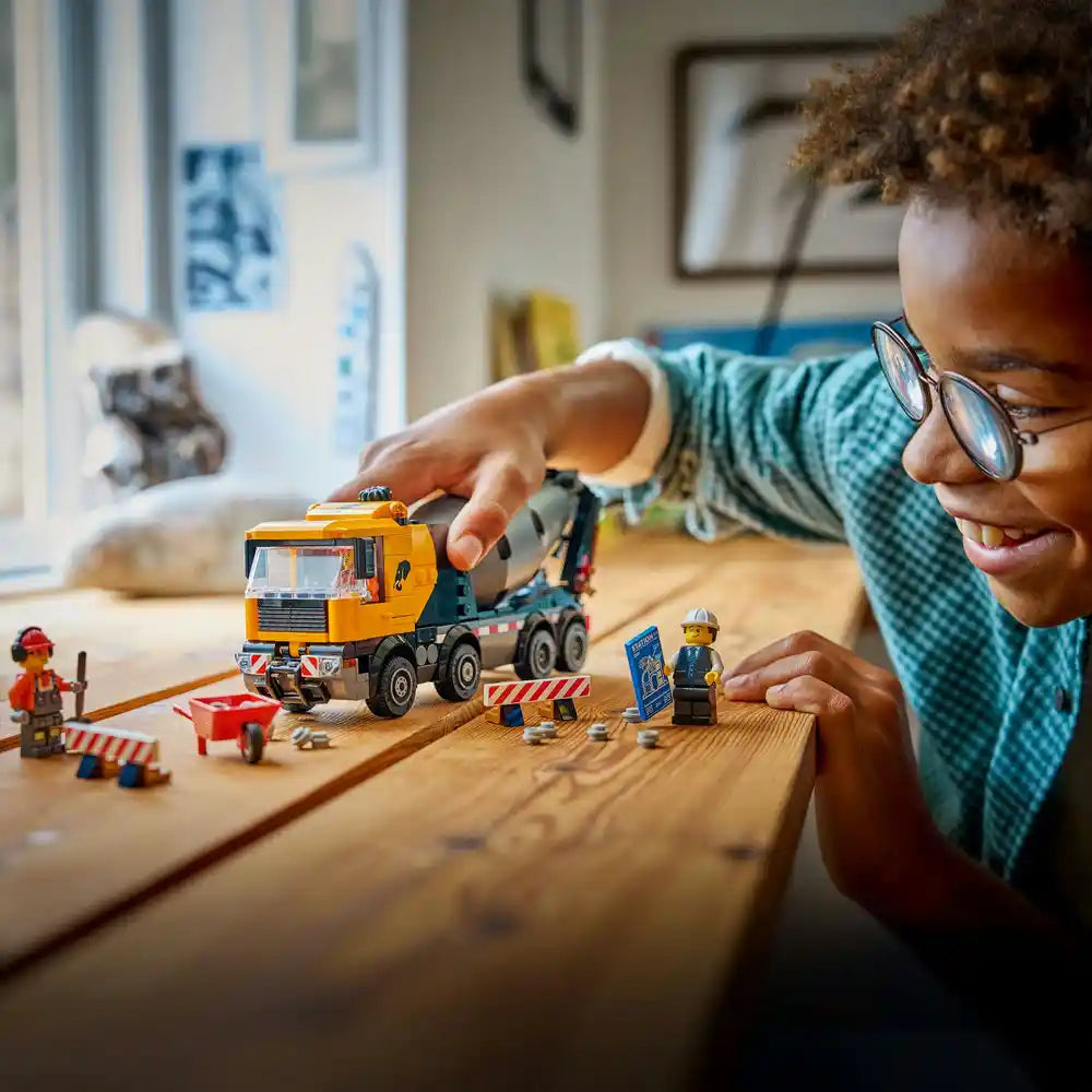 Child wearing glasses playing with LEGO City Cement Mixer set 60478, featuring an orange and black cement truck with construction minifigures and red and white barrier accessories on a wooden table
