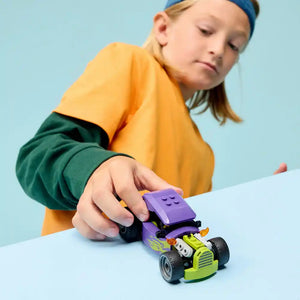 Child in yellow and green outfit playing with LEGO Hot Rod 60485 featuring a purple and lime green buildable race car with black wheels on white surface against light blue background
