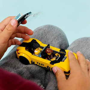 Child's hands holding a yellow LEGO taxi car set with minifigures on gray pants, with a LEGO glue pen visible above