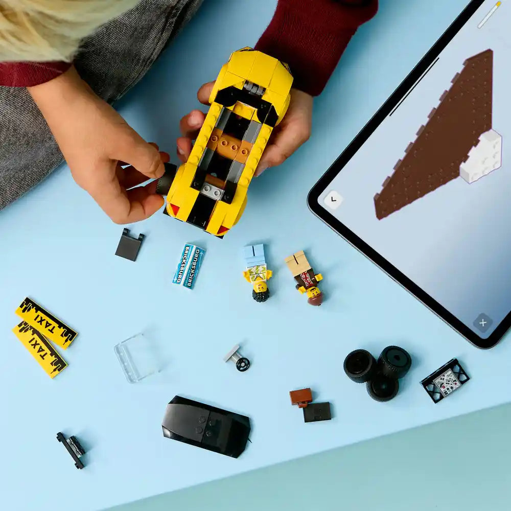 Child's hand holding a yellow and black LEGO taxi car with construction pieces, minifigures, batteries, wheels, and instruction manual displayed on light blue surface with tablet showing building guide