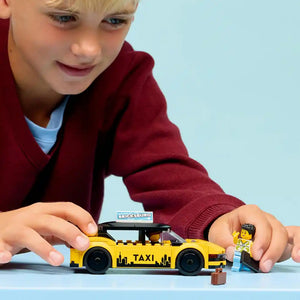 Young child in maroon sweater playing with LEGO yellow taxi set 60487, featuring bright yellow taxi car with black roof and LEGO minifigure driver against blue background