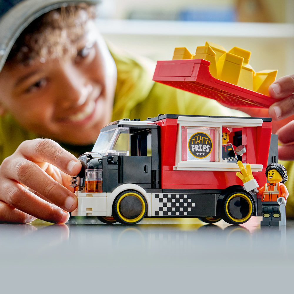 Child's hands playing with a LEGO food truck toy featuring red and black coloring with yellow fries on top, displaying a fries shop window and minifigure, on a light surface with blurred background