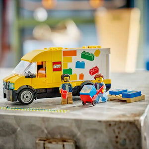 LEGO van set 60500 featuring a bright yellow and white delivery truck with LEGO brick graphics, two minifigures, a red shopping cart, wooden pallet, and scattered colorful LEGO bricks on a concrete surface with blurred background