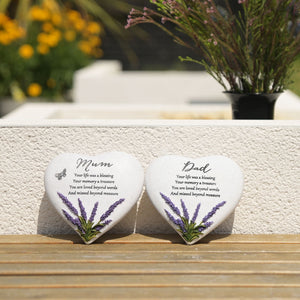 Two white heart-shaped memorial stones with lavender designs displayed on a wooden surface, one engraved for Mum and one for Dad, with potted flowers and foliage blurred in the background