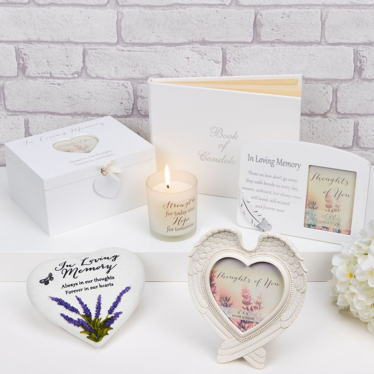 Collection of white sympathy and memorial gifts including a heart-shaped winged photo frame, decorative candle, Book of Condolences, heart-shaped stone with lavender design, and greeting cards arranged on white surface with flowers and white brick background