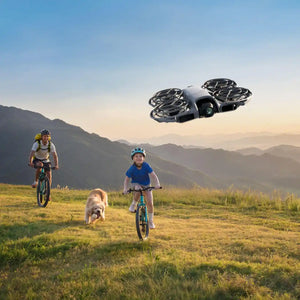 Futuristic white and black compact drone with four propeller guards hovering above a mountain landscape where a woman and child ride bicycles with a dog, representing aerial photography and outdoor adventure capabilities
