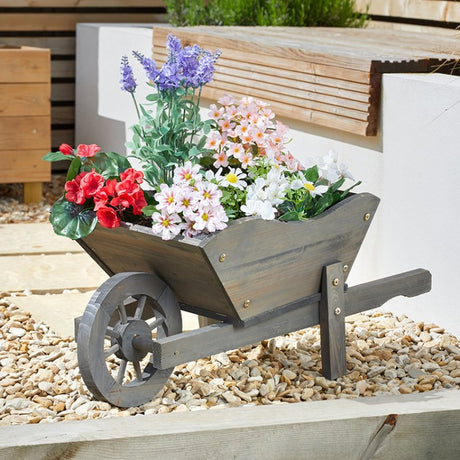 Wooden wheelbarrow planter with red, purple and white flowers in a garden setting on gravel, featuring a decorative wooden wheel and metal fixtures