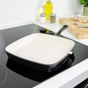 Blackmoor cast iron griddle pan with white ceramic cooking surface and black handle, displayed on induction cooktop in modern kitchen with cutting board, olive oil bottle, and fresh ingredients in background