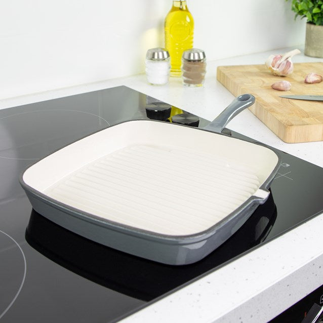Grey cast iron griddle pan with white ceramic interior on induction cooktop in modern kitchen with wooden chopping board, oil bottle, and salt and pepper shakers