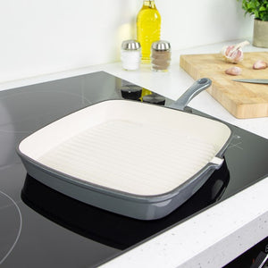 Grey cast iron griddle pan with white ceramic interior on induction cooktop in modern kitchen with wooden chopping board, oil bottle, and salt and pepper shakers