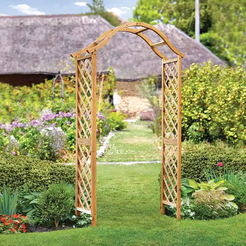 Wooden garden arch with lattice panels and curved top in tan finish, positioned in lush green garden with flowering plants and hedges, traditional countryside setting with thatched roof building in background