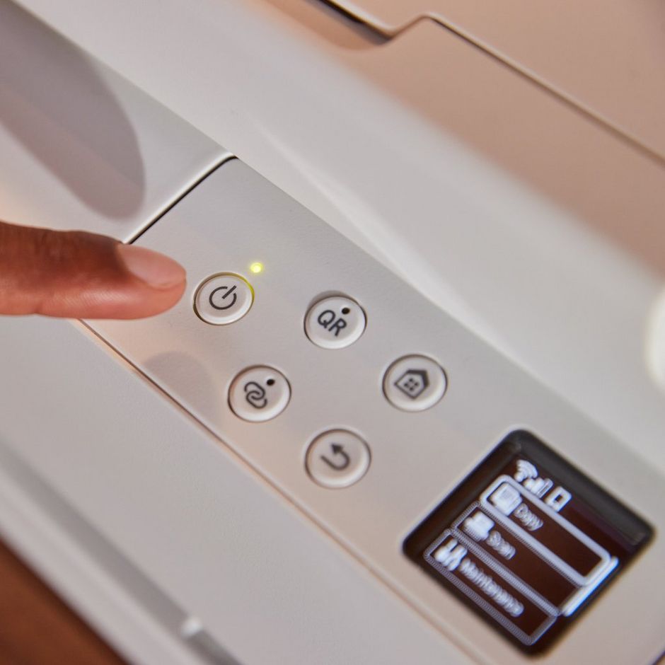 Close-up of Canon TS6550i white wireless inkjet printer control panel showing finger pressing power button with illuminated indicator light, circular function buttons for WiFi and copying, and paper tray slot visible on right side