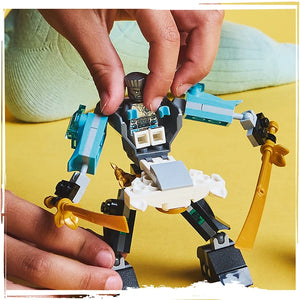 Child's hands holding a blue, white, black and gold LEGO Ninjago Zane's Battle Suit Mech figure against a bright yellow background with white socks visible