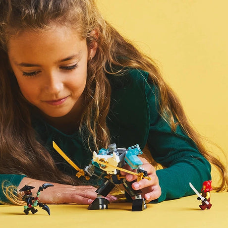 Young girl in dark teal long-sleeved shirt playing with LEGO Zane's Battle Suit Mech set against yellow background, displaying black and gold mecha figure with smaller minifigures