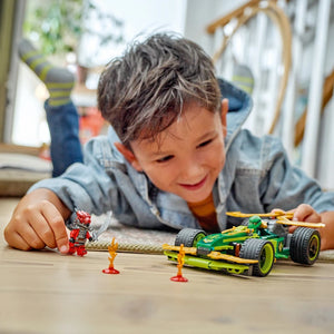 Young child playing with green and yellow LEGO pull-back race car set featuring Lloyd minifigure and orange traffic cones on wooden floor indoors