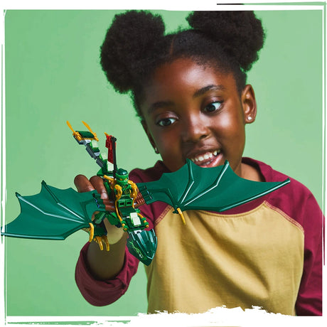 Young child with two puffs hairstyle holding and smiling at LEGO Lloyd's Green Forest Dragon set 71829, featuring a green and yellow dragon toy with wings against a green background, child wearing maroon and yellow color-block shirt