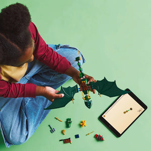 Child wearing red and yellow long-sleeved shirt and blue denim holding green and gold LEGO Lloyd's Green Forest Dragon set with black bat-like wings against mint green background with scattered LEGO pieces and instruction manual