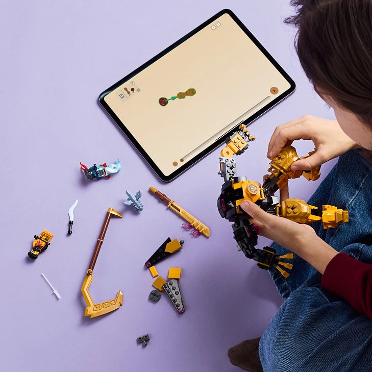Child assembling yellow and black LEGO Spinjitzu Battle Mech with tablet displaying building instructions on purple background, surrounded by individual LEGO pieces and components