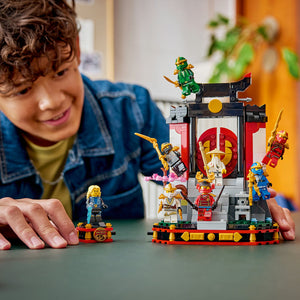 Child playing with LEGO Ninjago 15th Anniversary set featuring red and black display platform with golden ninja minifigures, yellow and red accents, and multiple character pieces on green surface