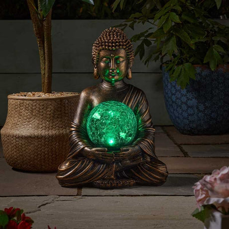 Bronze Buddha statue holding illuminated green glowing orb surrounded by potted plants and flowers in a garden setting at night