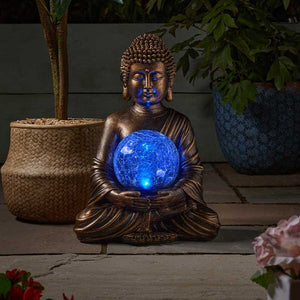 Bronze Buddha statue holding illuminated blue glowing orb sphere, seated in meditation pose surrounded by potted plants and flowers on wooden deck garden setting
