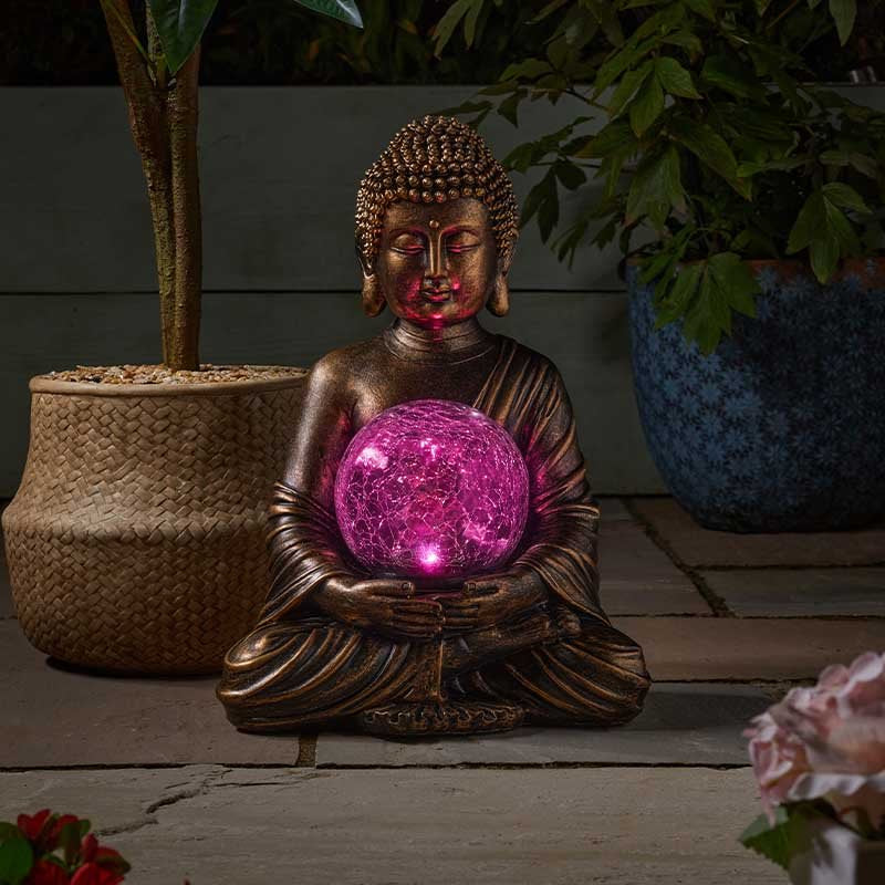 Bronze Buddha statue holding illuminated magenta LED crystal ball surrounded by potted plants and flowers on garden patio