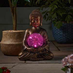 Bronze Buddha statue holding illuminated magenta LED crystal ball surrounded by potted plants and flowers on garden patio