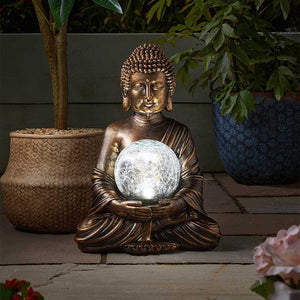 Bronze Buddha statue holding illuminated crackle glass solar light sphere, seated in meditation pose surrounded by potted plants and flowers in a garden setting