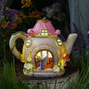 Whimsical fairy teapot solar light with pink curved roof, cream ceramic body, illuminated yellow windows and doors, decorated with colorful flowers and a tiny fairy figurine, surrounded by purple flowers and green plants in a garden setting