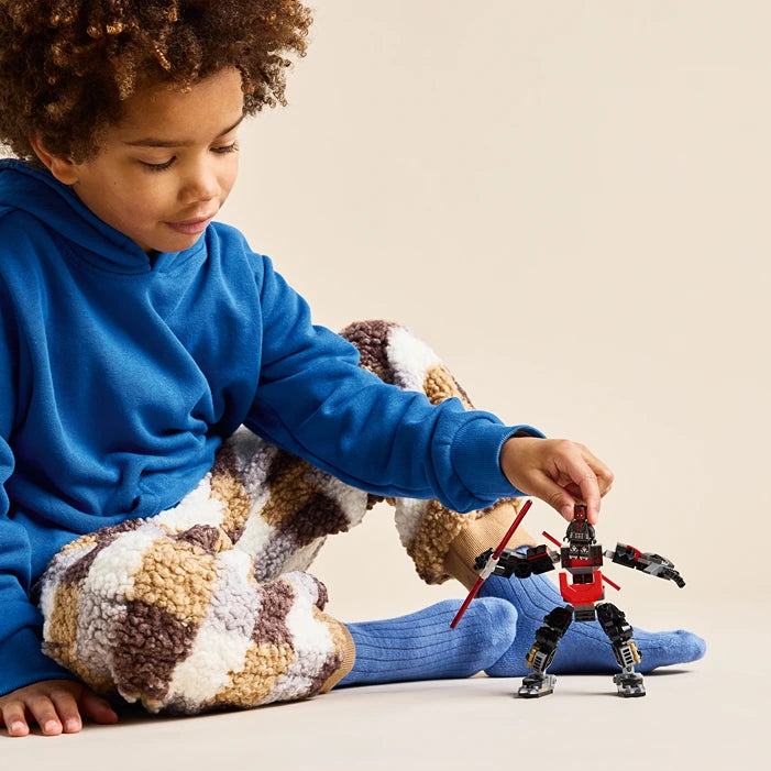 Young child in blue hoodie and blue socks playing with LEGO Darth Maul Mech set 75411, a red and black buildable robot figure, against neutral background