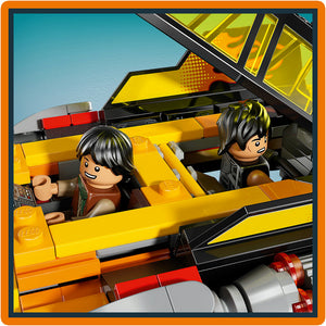 LEGO minifigures with black hair seated in a bright yellow and orange brick-built vehicle with black accents, featuring an open cockpit design and red and silver engine components visible below, set against a clear blue sky background