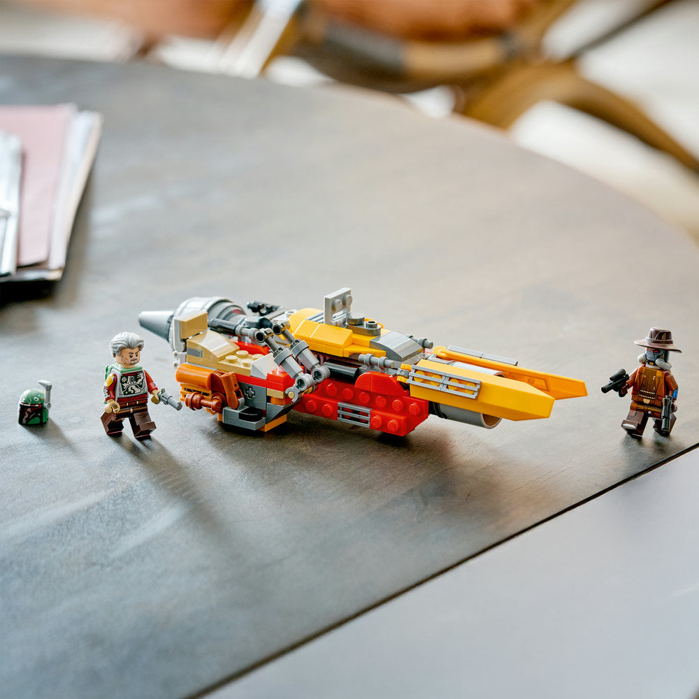 LEGO Cobb Vanth's Speeder set 75437 displayed on gray surface featuring orange and yellow spacecraft with red base, accompanied by four minifigures including Boba Fett character, against blurred background of building materials and wooden accessories