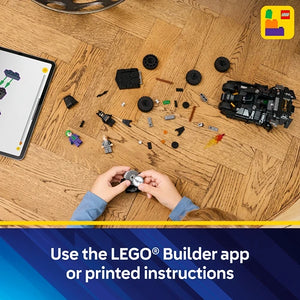 Child's hands assembling LEGO Batman Tumbler set on wooden floor surrounded by black and gray LEGO pieces, instruction manual, and building guide app interface displayed on tablet, with LEGO logo visible in corner