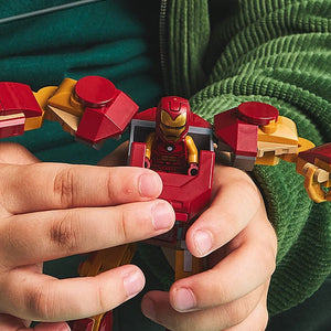 Child's hands holding LEGO Iron Man mech suit minifigure in red and gold with green knitted fabric and teal background visible alongside other red and yellow LEGO mech pieces from set 76307