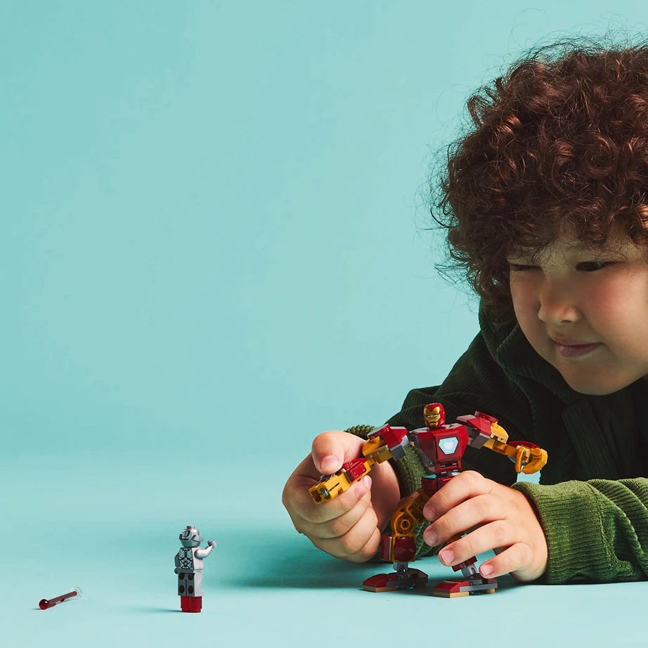 Child with curly brown hair playing with LEGO Iron Man Mech set 76307 featuring red and gold armored mech suit against turquoise blue background, with minifigures and building pieces visible