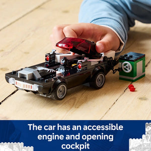 Child's hand holding red transparent hood above black LEGO muscle car with minifigures and green engine block on wooden surface, demonstrating removable hood feature