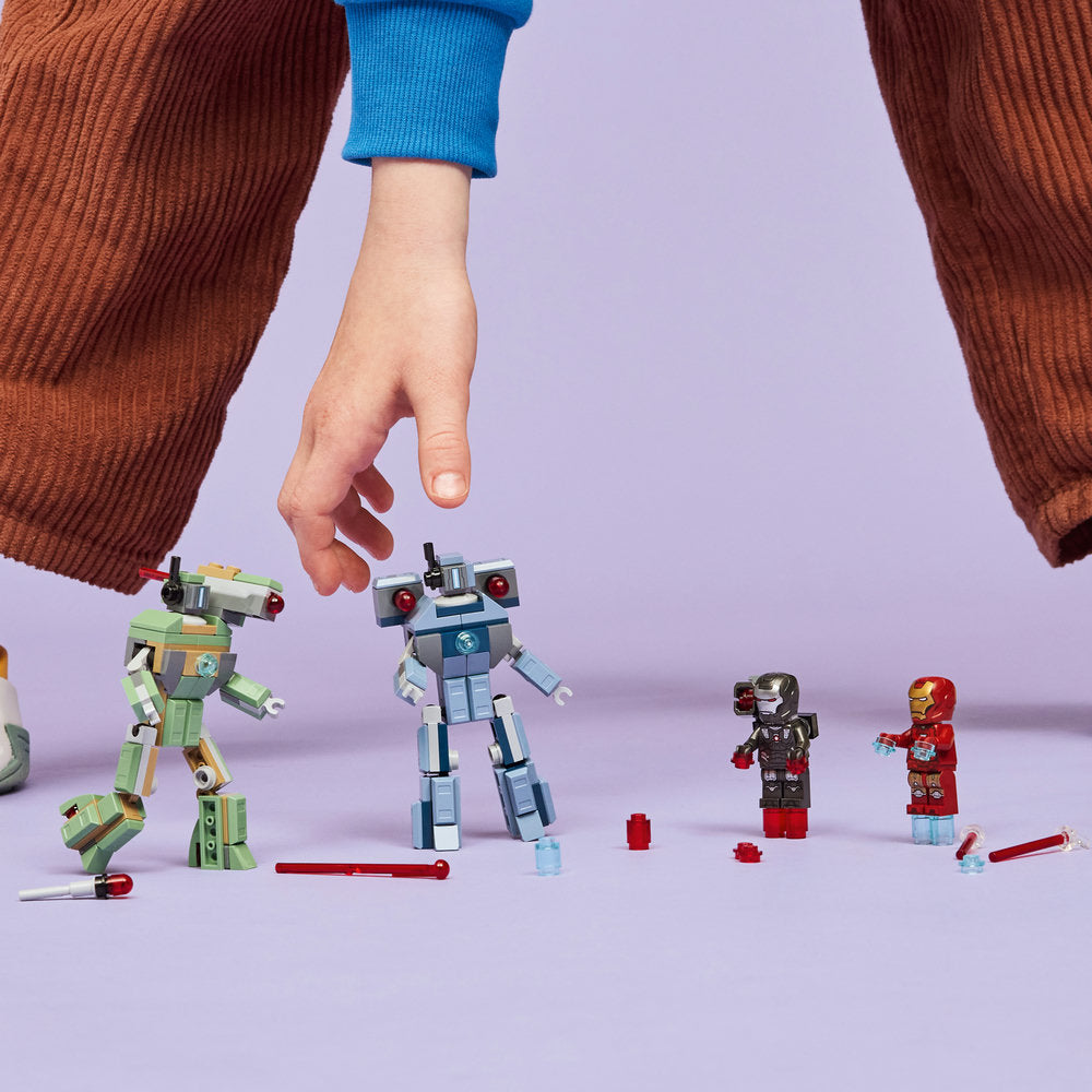 Hand holding LEGO minifigures above purple surface showing green and white robot, blue and white robot, Iron Man in black and gold, and War Machine in red and gold, with red LEGO pieces and accessories scattered nearby, wearing brown and blue knitted sweaters