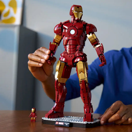 Large LEGO Iron Man Mark 3 buildable figure in red and gold displayed on a gray baseplate next to a small minifigure, held by a person's hand against a blurred background with blue clothing visible