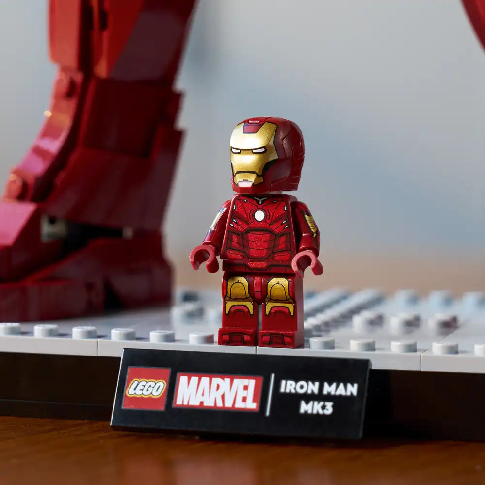 LEGO Marvel Iron Man MK3 minifigure in red and gold armor displayed on white baseplate with printed nameplate, featuring detailed chest arc reactor and helmet design, positioned in front of blurred red armor backdrop