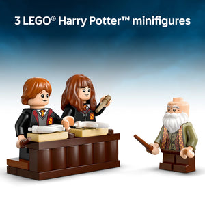 Three LEGO Harry Potter minifigures displayed against a blue gradient background featuring two students with reddish-brown hair in black Hogwarts robes with red and gold trim standing at a brown wooden desk with books and scrolls, and an elderly wizard with a white beard wearing olive green robes with a patterned vest holding a wand