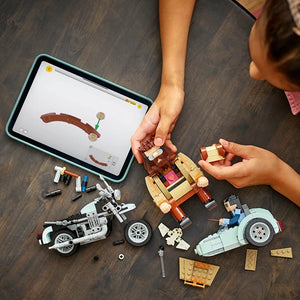 Child's hands assembling LEGO Hagrid's Motorcycle Ride set 76443 featuring brown motorcycle, tan and red brick structures, and white motorcycle models on wooden floor with instruction tablet displaying building schematic