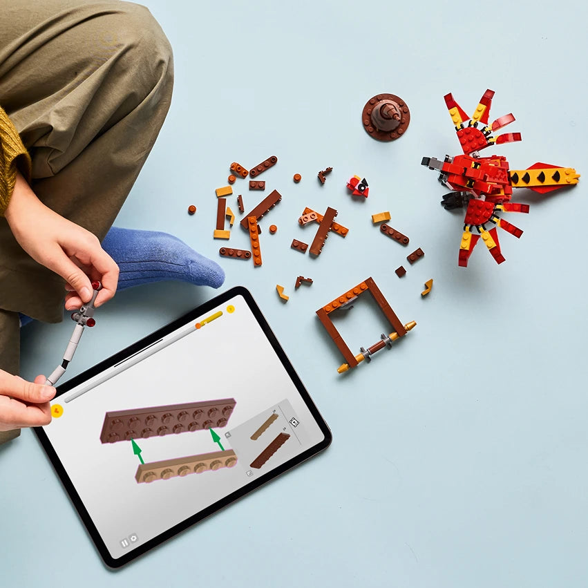 Child holding tablet displaying LEGO building instructions next to assembled red and gold LEGO Fawkes phoenix figure and scattered brown LEGO bricks on light blue surface, showing construction process for LEGO set 76448