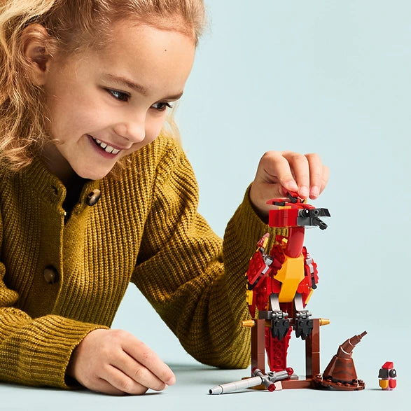 Child in mustard yellow cardigan playing with LEGO Fawkes phoenix set 76448, featuring red and gold buildable phoenix figure with black wings and brown perch against light blue background