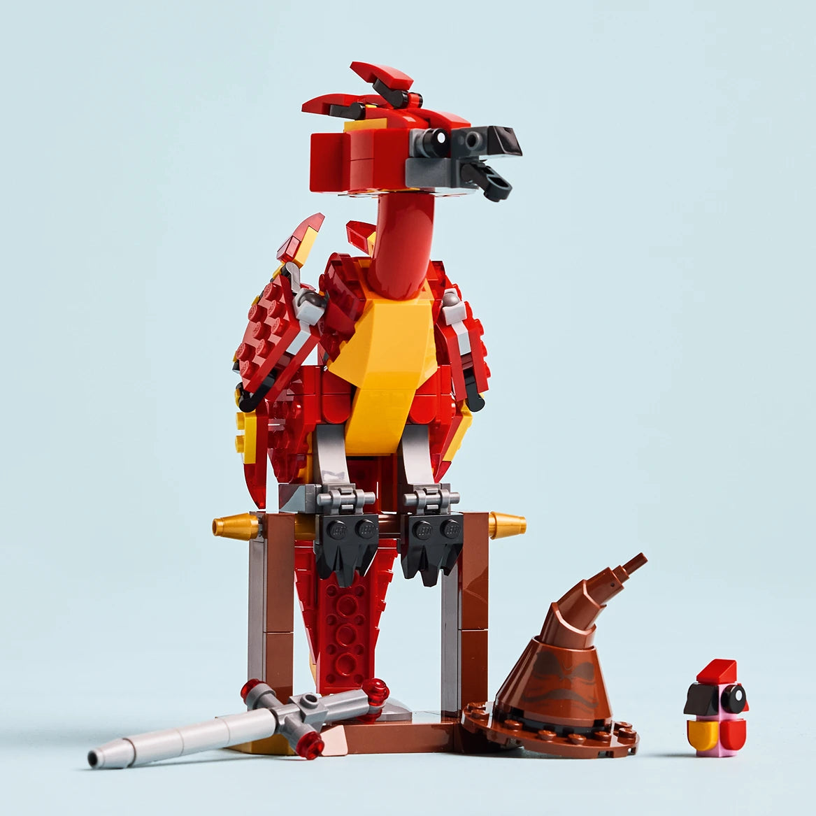 LEGO Fawkes Phoenix construction set 76448 featuring a red and yellow brick-built phoenix figure with articulated wings and head, mounted on a brown perch with golden accents, displayed with loose LEGO pieces and a minifigure component on light blue background