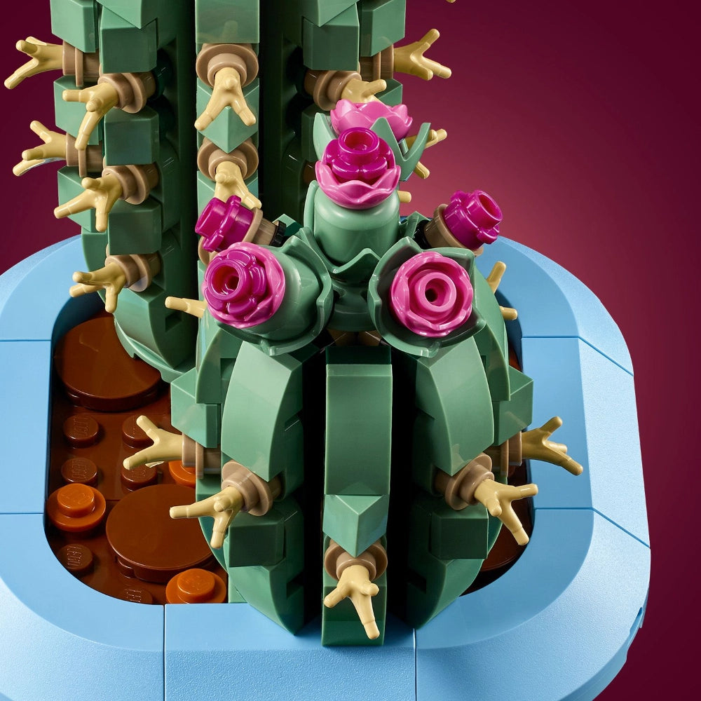 LEGO Flowering Cactus set 10309 featuring a green brick construction cactus with bright magenta pink flowers, tan and brown articulated arms, displayed on a light blue base plate against a deep burgundy background